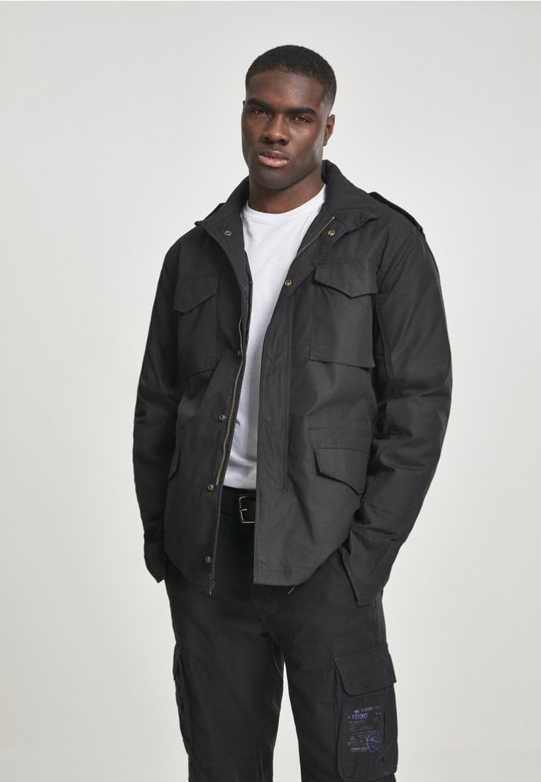 Black utility jacket with four front pockets, button closures, a zippered front, and adjustable cuffs, worn over a white T-shirt and black pants.