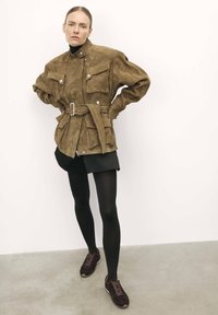 Brown suede shirt jacket with two chest pockets and a belted waist, paired with a black mini skirt and black tights, finished with dark sneakers.