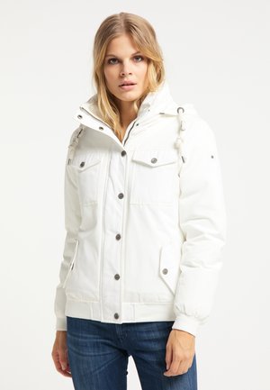 Femme blonde portant une doudoune blanche à capuche avec fermeture boutons et zip, et un jean bleu, debout devant un fond uni.