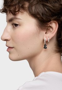 Jeune personne avec des cheveux bruns courts et bouclés portant des boucles d'oreilles en argent, dont une avec un petit pendentif en forme d'ours en peluche noir, sur un fond uni.