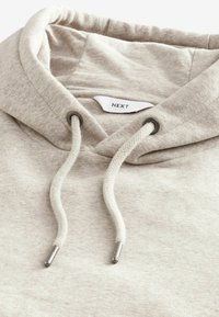 Sudadera beige con cordón y puntas metálicas plateadas, textura suave y una etiqueta con "NEXT" en la parte interior del cuello.
