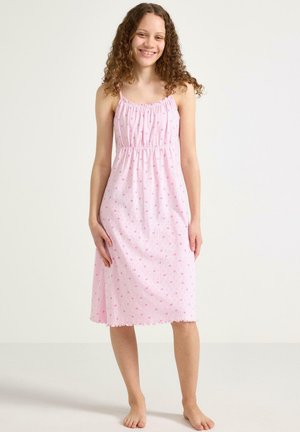 Fille souriante aux longs cheveux bouclés, portant une robe rose sans manches avec de petits motifs en forme de cœur, debout pieds nus devant un fond uni.