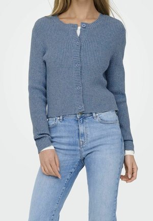 Femme portant un cardigan bleu côtelé boutonné sur un haut blanc, associé à un jean taille haute bleu clair sur un fond uni.