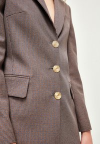 Brązowa blazer w kratę o dopasowanym kroju, z czterema złotymi guzikami i klapami. Materiał o teksturze oraz kieszenie boczne w komplecie.