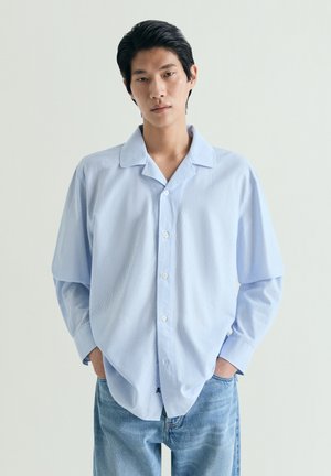 Joven con cabello corto y negro, que lleva una camisa de botones azul claro y jeans azules, de pie con las manos en los bolsillos frente a un fondo liso.