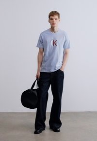 Ανοιχτό μπλε μπλουζάκι με λογότυπο, σε συνδυασμό με σκούρο μπλε τζιν παντελόνι με φαρδιά μπατζάκια. Στο αριστερό χέρι της φιγούρας κρατά μια μαύρη τσάντα τύπου duffel.