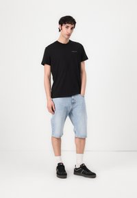 Schwarzes T-Shirt mit kurzen Ärmeln und kleinem Logo, kombiniert mit hellblauen Jeanshorts. Das Modell trägt schwarze Sneakers und weiße Socken.