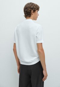 Polo a maniche corte in bianco, con un design a maglia texturizzata, colletto piatto e orlo a costine, indossato con pantaloni scuri. Vista da dietro.