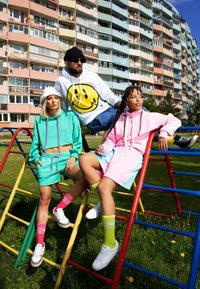 Trzy modele w kolorowych bluzach z kapturem: miętowozielonej, różowej i białej z uśmiechniętą buźką. Jasne skarpetki i białe sneakersy; w tle plac zabaw.