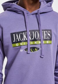 Lilla hettegenser med en svart og grønn grafikk som har teksten "JACK & JONES" og "ORIGINALS", snorer og en myk bomullstekstur.