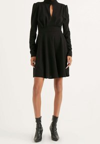 Robe noire avec un corsage ajusté, de longues manches bouffantes et un décolleté en forme de clé. Jupe évasée, confectionnée dans un tissu lisse. Bottines noires à la cheville.