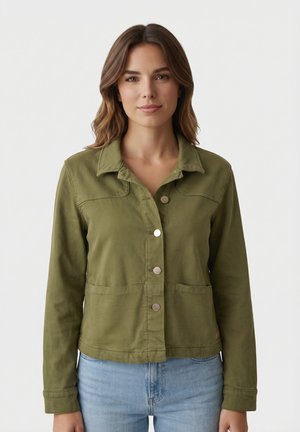 Donna con capelli castano medio fino alle spalle, indossa una giacca verde oliva con bottoni e jeans azzurri, in piedi davanti a uno sfondo bianco uniforme.