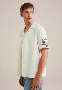 Camisa de botones de manga corta en tejido amarillo claro, con bordados florales en la manga izquierda. Llevada sobre una camiseta blanca sin mangas.