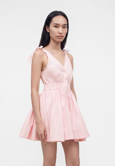 Γυναίκα με μακριά μαύρα μαλλιά που φοράει αμάνικο ανοιχτό ροζ φόρεμα με δεσίματα στους ώμους και σούρες στη μέση, στέκεται μπροστά σε μονόχρωμο φόντο.