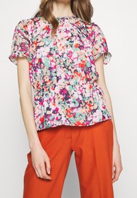 Blusa floral con escote fruncido, mangas cortas y cintura de peplum. Los colores incluyen rosa, verde y morado sobre un fondo claro.
