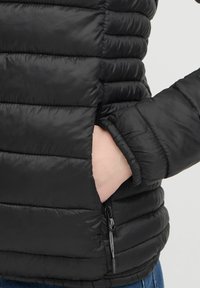 Čierna pérová bunda s prešívanými časťami, bočnou vreckom na zips a priliehajúcimi manžetami. Hladká textúra s elegantným dizajnom.