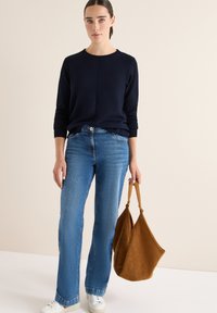 Marineblauer Strickpullover, hellblaue Schlagjeans und eine beigefarbene Wildledertasche. Weiße Sneakers runden das Outfit ab. Glatte Textur, lässiges Design.