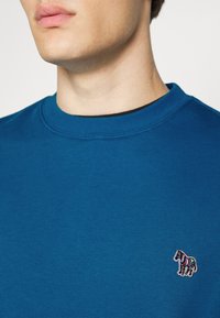 Blauwe crewneck trui gemaakt van zachte stof met een kleine multicolor zebraagwerk op de linkerborst.