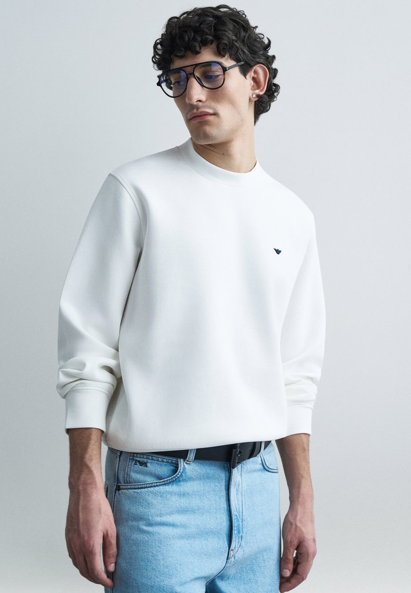 Λευκό φούτερ με στρογγυλή λαιμόκοψη και μακριά μ�ανίκια, με μικρό μαύρο λογότυπο στο στήθος. Φορεμένο με ανοιχτόχρωμα μπλε τζιν.