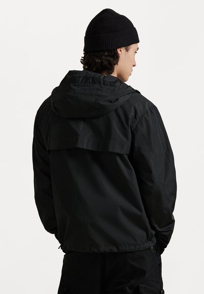 Μαύρο εξωτερικό μπουφάν με κουκούλα, featuring σχεδίαση με επίπεδα στο πίσω μέρος και ελαφριά, λεία υφή. Φορεμένο με μαύρη πλεκτή μπερέ.