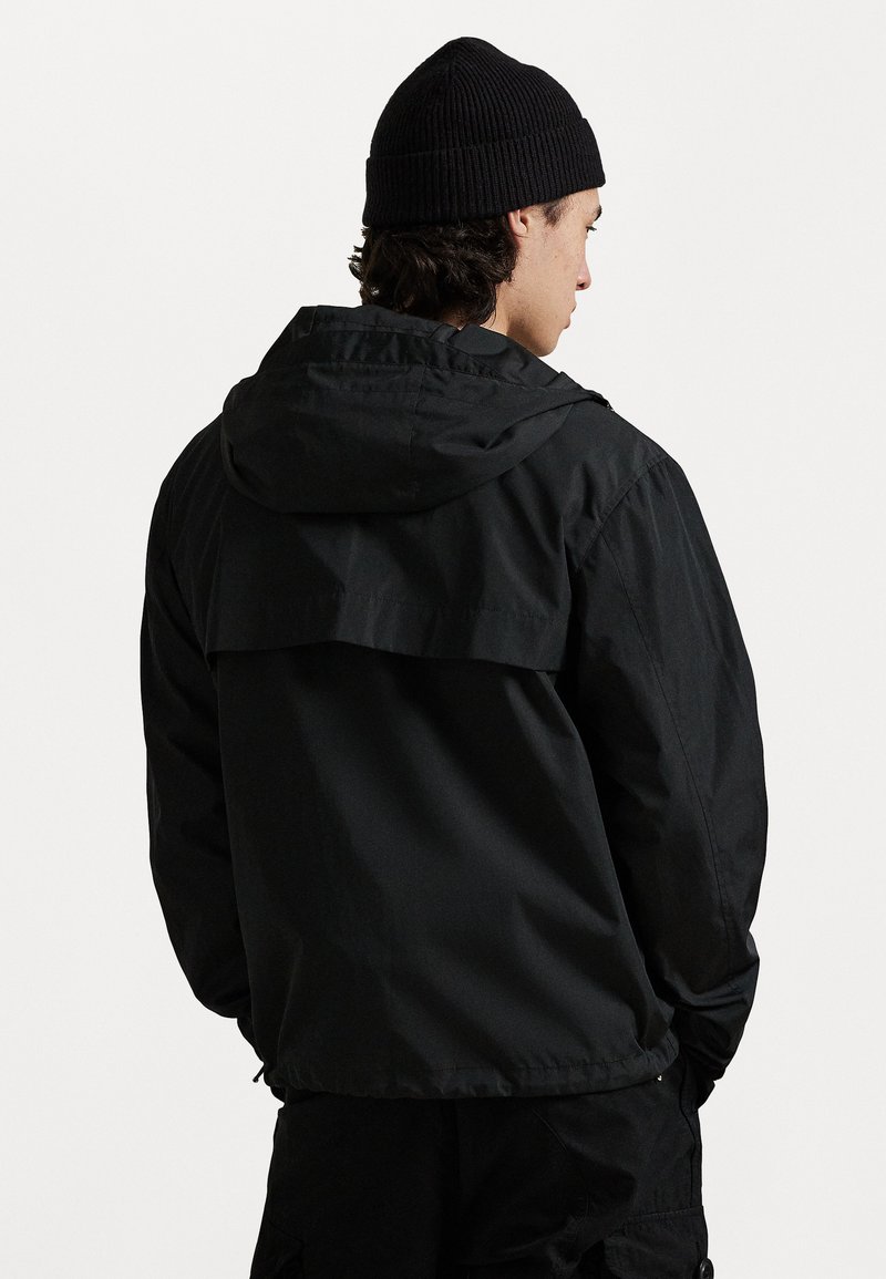 Veste extérieure noire avec capuche, dotée d'un design à dos superposé et d'une texture légère et lisse. Portée avec un bonnet en tricot noir assorti.