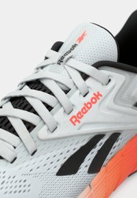 Pantofii atletici Reebok de culoare gri deschis au un superior din plasă texturată, accente negre, o talpă portocalie și detalii proeminente ale logo-ului.