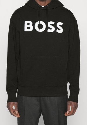 Henkilö pukeutuneena mustaan huppariin, jossa valkoinen "BOSS"-teksti rinnassa, yhdistettynä tummanharmaisiin housuihin, seisoo yksiväristä taustaa vasten.