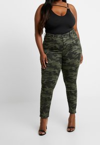 Mujer vestida con jeans ajustados de camuflaje verde, top sin mangas negro y tacones abiertos negros, de pie contra un fondo blanco liso.