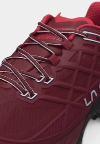 Primo piano di una scarpa da atletica rossa con lacci dettagliati, tomaia testurizzata e logo "LA" branded sul lato.