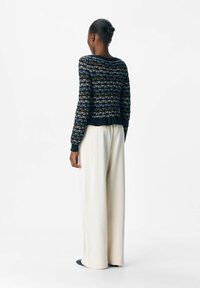 Pull noir à motifs géométriques en bleu et or, avec un ourlet côtelé, associé à un pantalon large blanc et des chaussures noires.