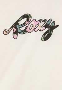 Un tissu en coton blanc arbore le mot "Roxy" dans une calligraphie stylisée. Les lettres sont remplies de motifs floraux dans des teintes de rose, vert et noir.