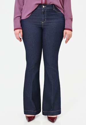 Donna che indossa jeans a zampa blu scuro, una blusa viola a maniche lunghe e tacchi a punta color bordeaux, in piedi su uno sfondo bianco.