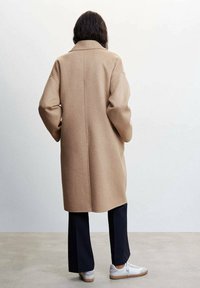 Abrigo de lana beige con cuello, de corte oversize, hombros caídos y silueta recta, combinado con pantalones negros y zapatillas blancas.