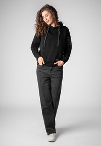 Zwarte capuchontrui van textielstof met grijze trekkoorden; gecombineerd met losse, donkergrijze jeans en witte sneakers.