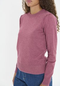 Suéter de punto en rosa suave con cuello redondo, puños y dobladillo acanalados. Presenta mangas abullonadas para un detalle adicional. Llevado con jeans azules.