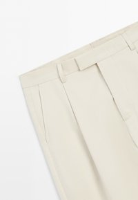 Light beige tailored trousers with belt loops, side pockets, and a pleated front, displayed on a white background.