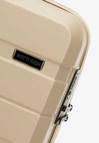 Valigia rigida beige con finitura testurizzata, dotata di una placca con logo nera e doppie cerniere sul lato. Design elegante e moderno.