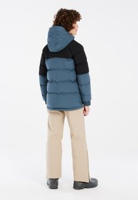 Giacca imbottita con blocchi di colore blu e nero, dotata di cappuccio, polsini elasticizzati e di un design a lunghezza vita, indossata con pantaloni beige e stivali neri.