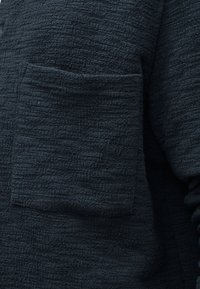 Tissu foncé texturé avec une poche carrée sur la poitrine d'un vêtement à manches longues, montrant un tissage détaillé et des nuances subtiles.