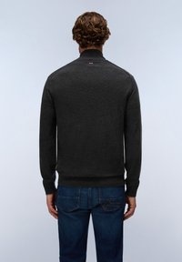 Man with short curly hair wearing dark gray sweater and blue jeans, standing with his back to the camera against a plain light background.