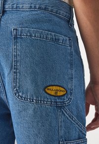 Bermudas cargo de mezclilla azul con un bolsillo de parche que presenta una etiqueta amarilla de "Billabong". Tejido texturizado con costuras visibles y un ajuste holgado.