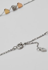Sølv kædearmbånd med tre hjertecharms i gult metal, sølv og rosaguld. Inkluderer en rund lås og en forlængerkæde.