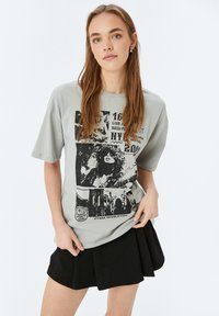 Lichtgrijze oversized t-shirt met zwarte grafische prints en tekst; korte mouwen; gecombineerd met een zwarte plooienrok.