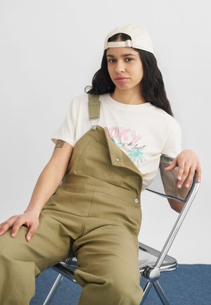 Νεαρός με μακριά σκούρα μαλλιά, φοράει μπεζ καπέλο με το μπροστινό μέρος προς τα πίσω, λευκό t-shirt με γραφικό σχέδιο και λαδί ολόσωμη φόρμα, καθισμένος σε μεταλλική αναδιπλούμενη καρέκλα.