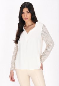 Top bianco con un corpo in tessuto morbido, scollatura a V e maniche lunghe con dettagli in paillettes argentate lucide. Texture liscia in tutto il capo.