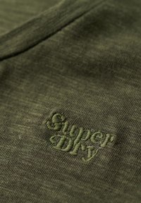 Tela de algodón verde oliva con un acabado texturizado; presenta el logo bordado "Super Dry" en hilo verde a juego.