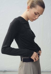 Črna dolgotrajna majica z drapiranim dizajnom in teksturirano tkanino, kombinirana s svetlo sivimi hlačami z visokim pasom in temnim pasom.