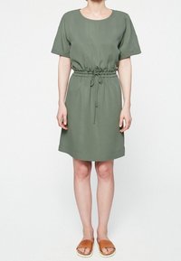 Robe verte à manches courtes avec une taille cintrée, un détail à cordon de serrage et une coupe droite, complétée par des sandales plates marron clair.