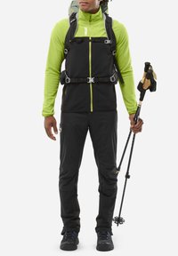 Black and green hiking outfit with a zippered top, adjustable straps, and black pants, holding trekking poles, suitable for outdoor activities.