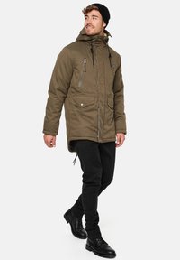 Parka verde măsliniu cu glugă, două buzunare frontale și buzunare cu fermoar pe piept. Pantaloni negri și bocanci negri completează ținuta.